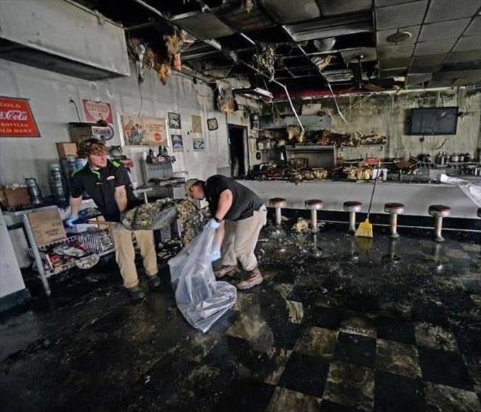 Two men with a shovel and a plastic bag cleaning debris inside a restaurant due to fire damage